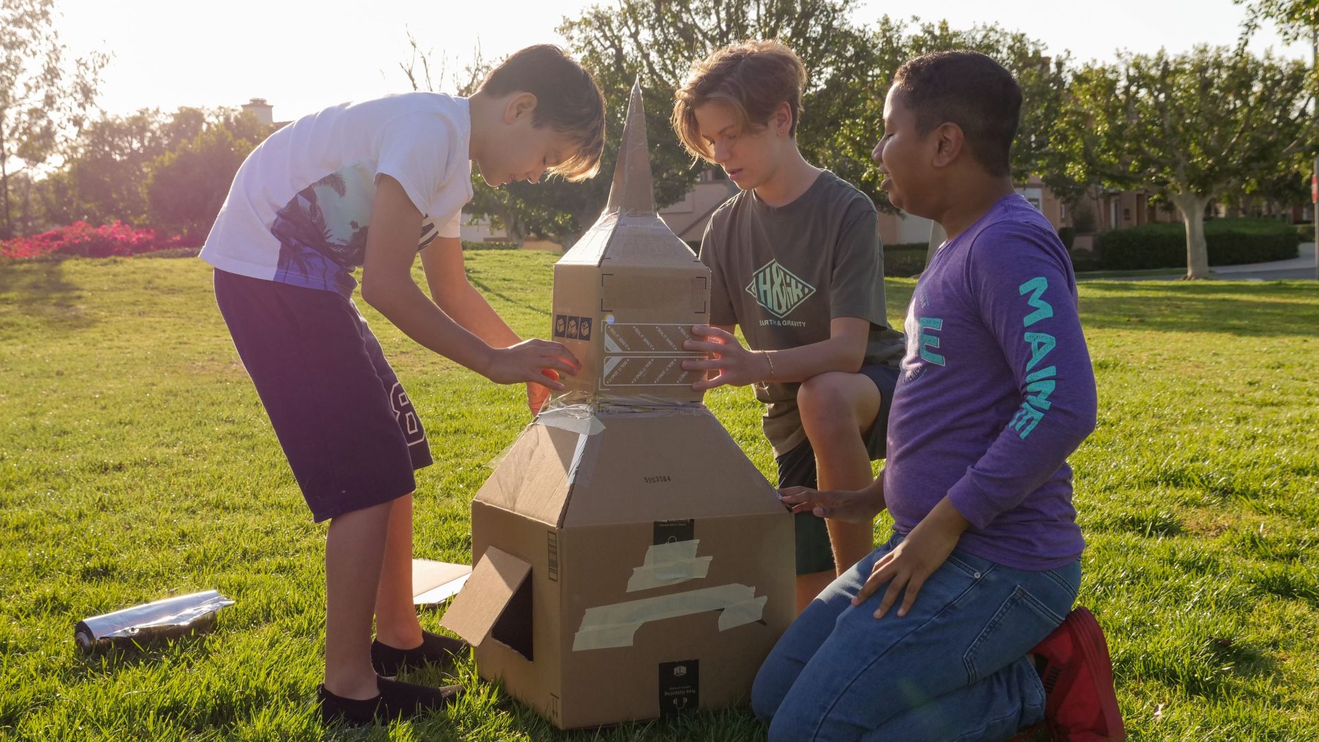 Three boys engaged in creative play, constructing a cardboard rocket outdoors.
